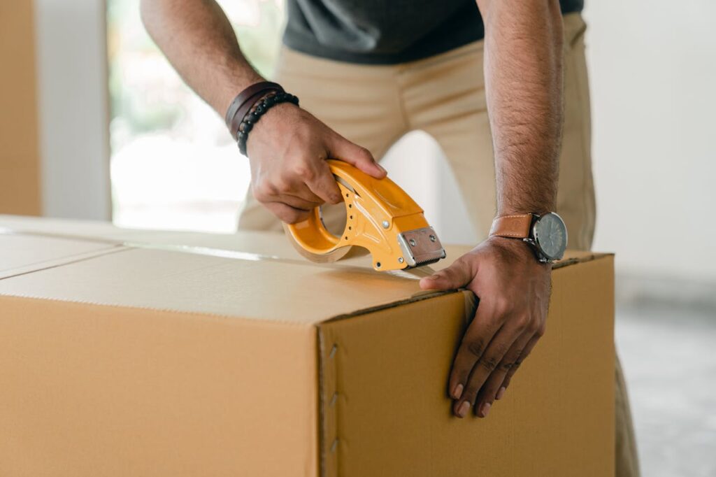 pexels-photo-4246120-1 Crop faceless young male with wristwatch using adhesive tape while preparing cardboard box for transportation